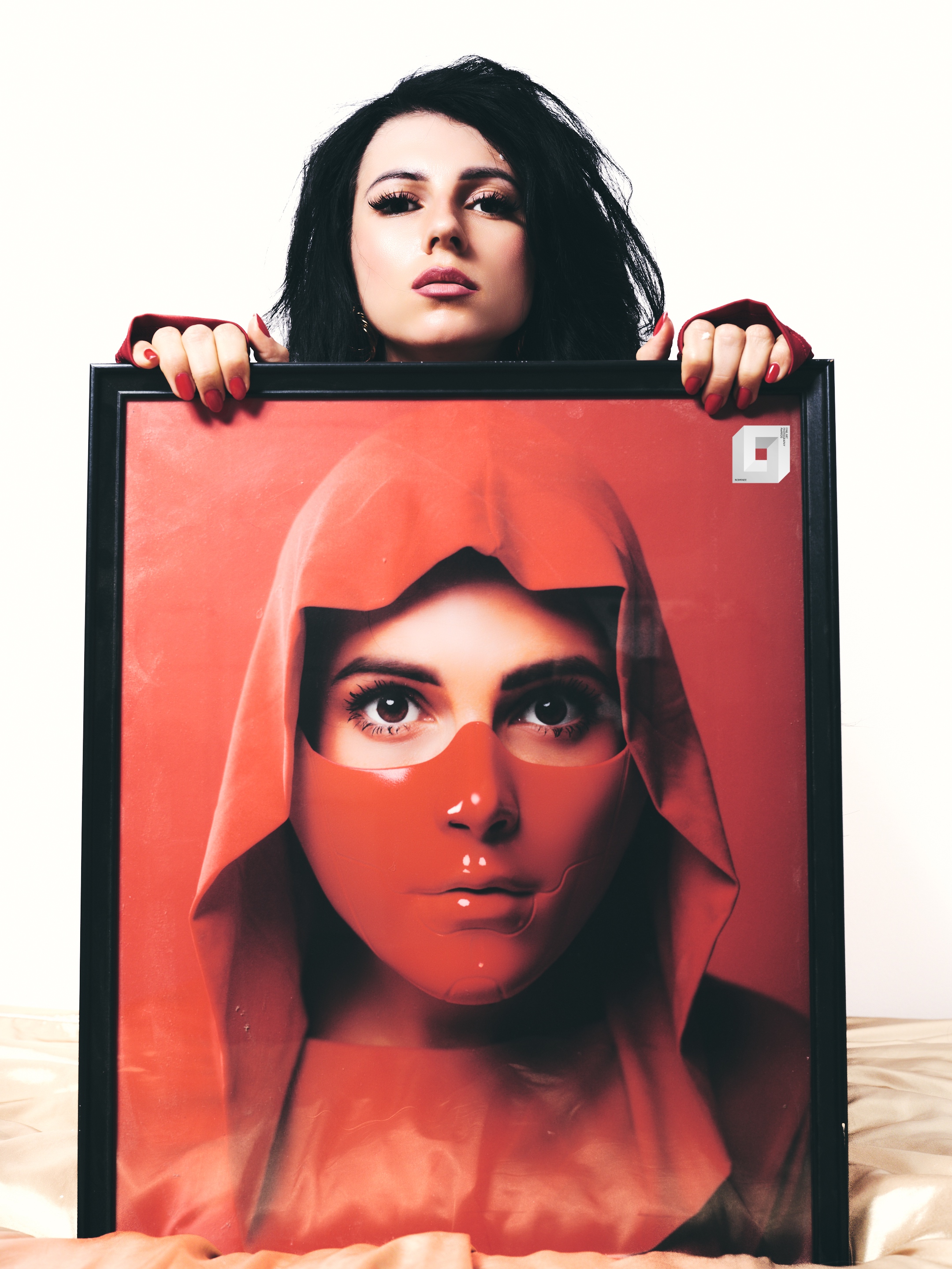 A square-formatted artwork held in a black frame by a person positioned behind it against a plain white background. Only the person's upper face—dark hair, dark eyebrows, and intense brown eyes gazing directly forward—and her hands with red-painted fingernails resting on the top edge of the black frame are visible above the picture. Inside the black frame is a photo of a woman against a deep red background. She wears a bright red hood framing her head and a glossy, sculpted red lower-face mask that covers her mouth, chin, and nose, leaving only her upper face visible. Her dark eyes stare straight ahead, illuminated by soft front lighting with subtle highlights reflecting off the shiny mask surface. A small square logo sits in the top-right corner of the framed picture. The composition features strong geometric lines from the frame contrast against the soft, organic shapes of the face, with a bold red, black, and white color scheme.