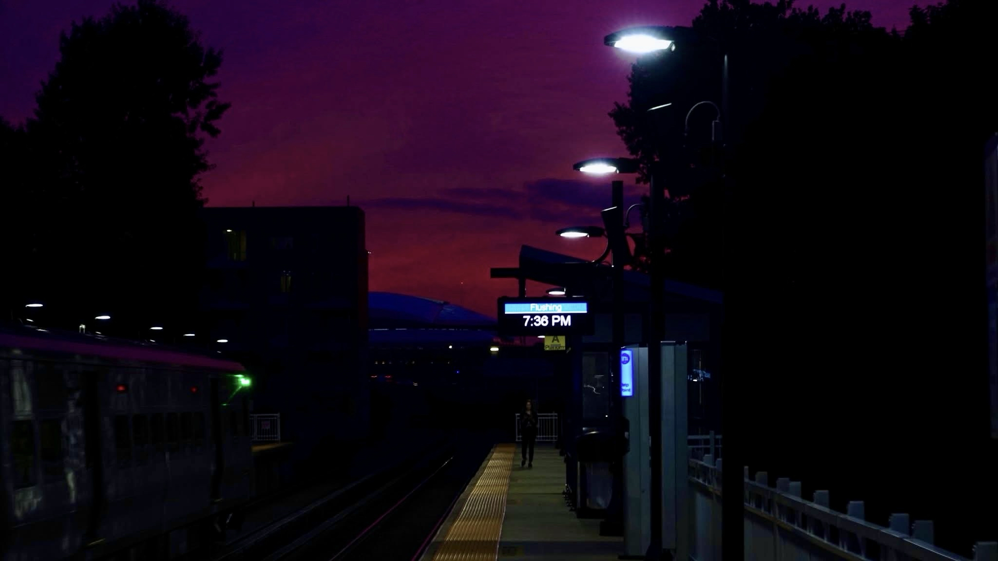 Photograph of the westbound
Platform of the Long Island railroad station at Main St., Flushing. Going down the lower center of the image is the platform with a yellow stripe on the left-hand side with one person towards the end of the platform. There are various lights. There’s a clock with the TrainTime above it and other train station accoutrements across the tracks. There is an eastbound train. You can see the outline of some buildings and some trees and the sky is magenta.