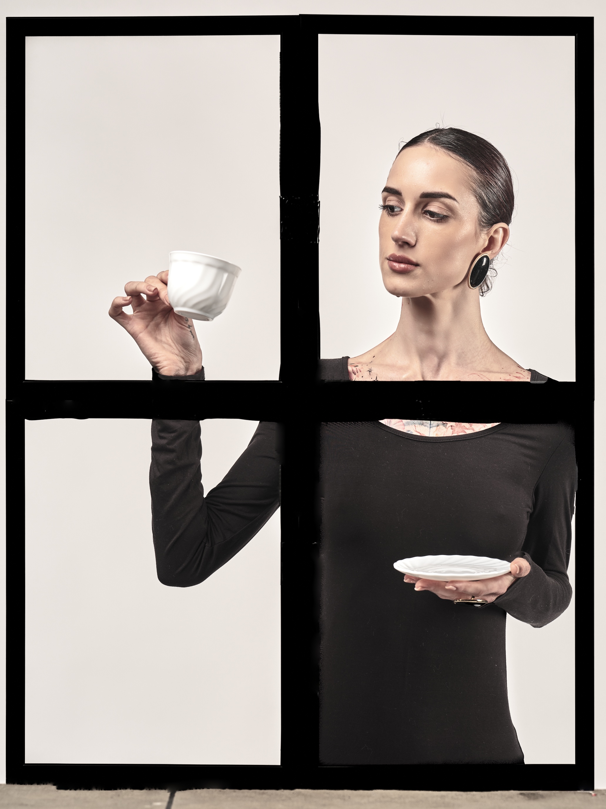 A portrait of a fair-skinned woman with dark, tightly pulled back hair, positioned behind a thick, black cross-shaped frame that divides the composition into four quadrants against a flat off-white background. The woman occupies the right half and center. She wears a form-fitting, long-sleeved black top with a scoop neckline revealing faint dark tattoos on her upper chest. She has large, flat, circular black earrings with thin gold rims. Her head is turned left, gaze directed downward at a stark white, fluted teacup in her elevated right hand within the top-left quadrant. Her left hand, in the bottom-right quadrant, holds a textured white saucer flat at waist level, displaying a dark ring. The black frame bisecting the image has slightly irregular edges, resembling thick tape or painted lines rather than rigid architecture. Lighting is soft yet directional from the upper left, creating gentle shadows under her jawline, nose, and beneath the teacup, while the background remains evenly i