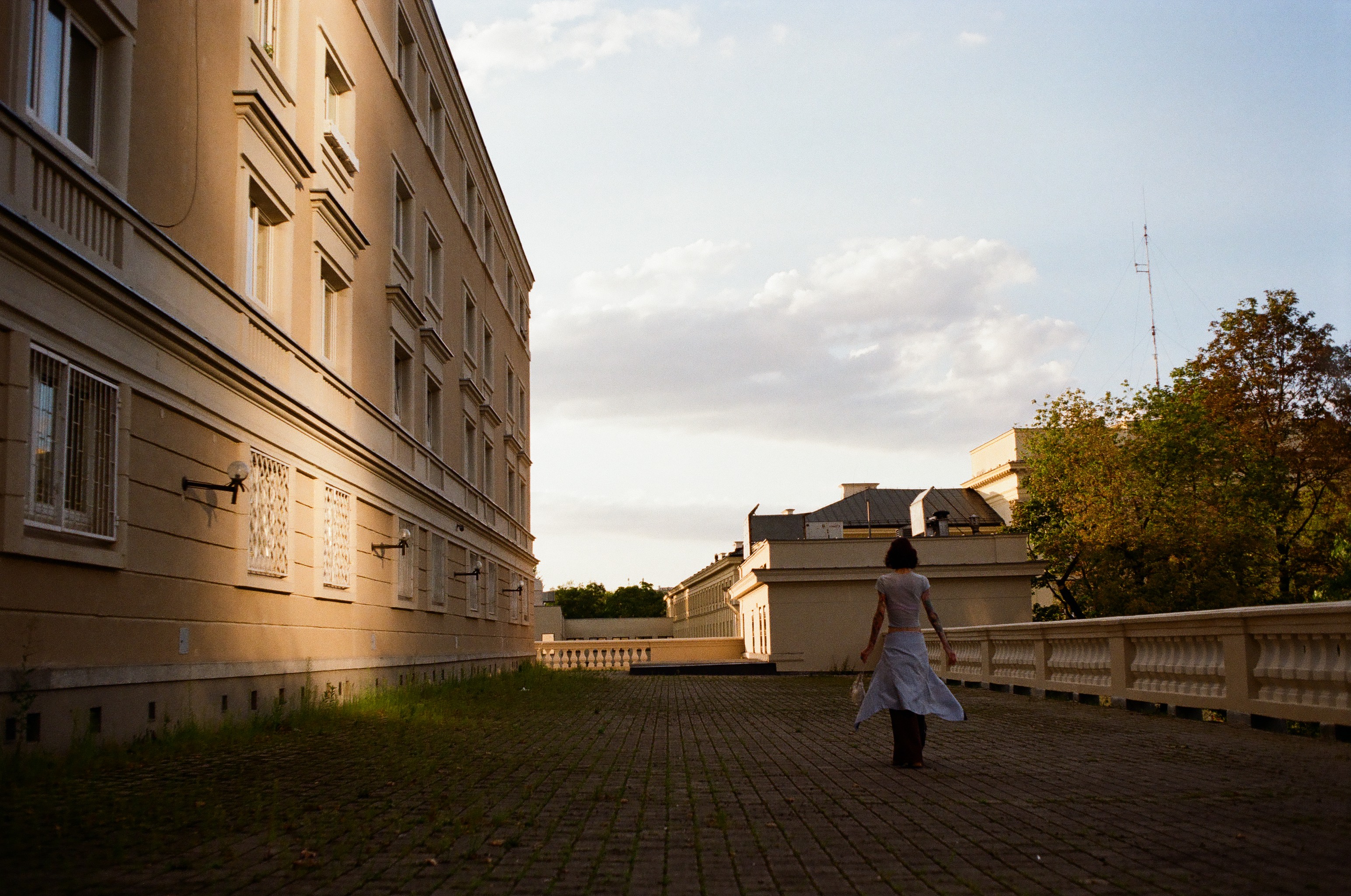 A wide, eye-level photograph taken outdoors during late afternoon light on a stone-paved terrace. On the left side of the frame stands a large, multi-story beige building with a textured facade, square windows, and horizontal moldings casting long shadows. Warm, golden sunlight hits the upper facade and the ground, while cool shadows cover the lower left section where sparse grass grows between paving stones. On the right, a low balustrade with decorative vertical balusters runs parallel to the building, leading the eye toward the background. A person with dark, short hair is seen from behind, walking away from the camera in the right-center foreground. They wear a light-colored top, dark pants, and a light shirt or jacket tied loosely around their waist, flowing behind them as they move. In the background, lower cream-colored building structures, lush green trees, and an antenna pole are visible against a soft sky filled with light, scattered white clouds illuminated by the setting su